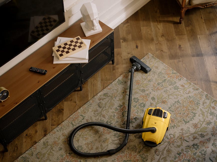 A yellow canister vacuum cleaner with a black hose and extension wand is positioned on a patterned rug in a living room with wooden floorboards. To the left, a black metal and wood console table holds a remote control, a stack of white papers, a chessboard, and a white decorative bust. The room features warm lighting, and seen in the background is a part of an armchair with wooden armrests and a dark fabric cushion. The scene illustrates indoor surface cleaning by Chiswick Carpet Cleaning, focusing on maintaining hygiene and cleanliness in a domestic setting.