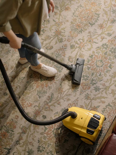 A person using a vacuum cleaner with a black hose attachment to clean a patterned, beige floral carpet in a residential setting. The individual is wearing beige shoes and grey jeans, standing on the carpet, which is part of a living room or lounge area with soft lighting. The vacuum cleaner is yellow with black accents and is positioned on the carpet, connected to a power outlet nearby. The carpet appears clean and well-maintained, with visible floral motifs and a mix of muted colors. The scene reflects surface cleaning and maintenance by Chiswick Carpet Cleaning, emphasizing deep cleaning and hygiene of household flooring surfaces.