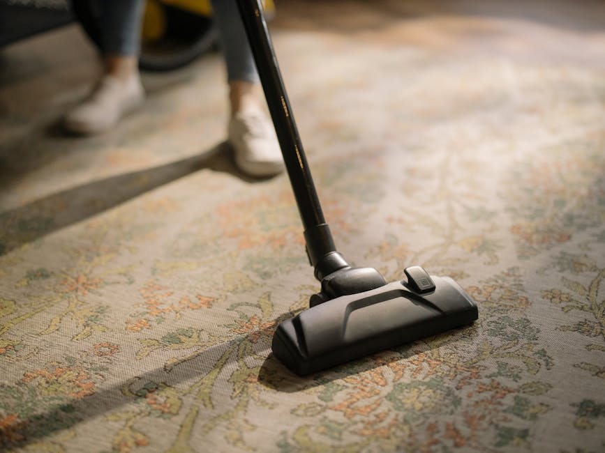 A yellow canister vacuum cleaner with a black hose and extension wand is positioned on a patterned rug in a living room with wooden floorboards. To the left, a black metal and wood console table holds a remote control, a stack of white papers, a chessboard, and a white decorative bust. The room features warm lighting, and seen in the background is a part of an armchair with wooden armrests and a dark fabric cushion. The scene illustrates indoor surface cleaning by Chiswick Carpet Cleaning, focusing on maintaining hygiene and cleanliness in a domestic setting.