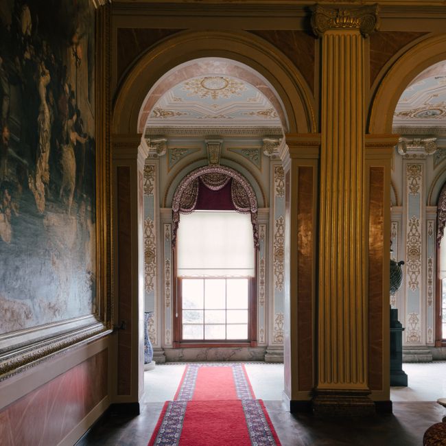 Interior view of a grand, ornately decorated room with high ceilings and classical architectural details, including tall, fluted gilded columns and intricate molding. A large window with a white roller blind allows natural light to illuminate the space, which features detailed painted ceiling decoration. The floor is partially visible with a red carpet runner leading towards the window, highlighting the room's elegant and historic design. The scene emphasizes the room's cleanliness and well-preserved condition, reflective of high standards in surface cleaning and maintenance, suitable for professional domestic or commercial cleaning services as offered by Chiswick Carpet Cleaning.