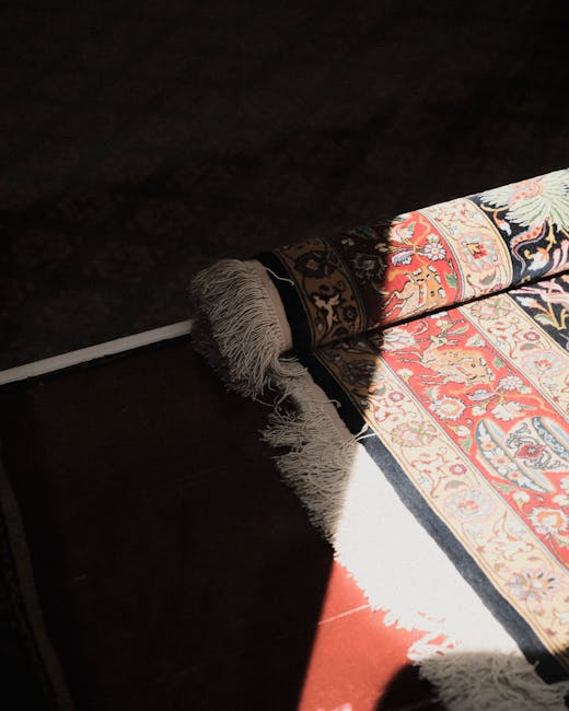 A section of a Persian-style rug with intricate floral and geometric patterns in red, blue, beige, and cream, partially unrolled on a dark wooden floor in a well-lit room near Chiswick House. The rolled edge reveals plush, fringed tassels, and sunlight creates a bright contrast between the carpet and the surrounding area. The scene suggests professional surface cleaning and deep carpet maintenance, aligning with services offered by Chiswick Carpet Cleaning in the Bedford Park area.