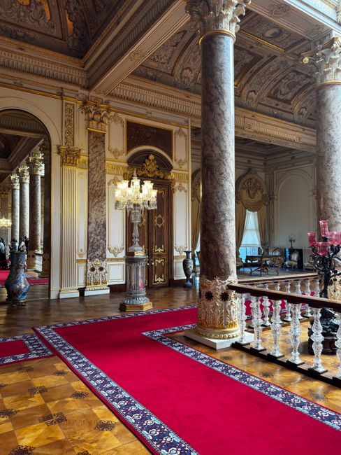 Interior view of a grand, ornately decorated room with high ceilings and classical architectural details, including tall, fluted gilded columns and intricate molding. A large window with a white roller blind allows natural light to illuminate the space, which features detailed painted ceiling decoration. The floor is partially visible with a red carpet runner leading towards the window, highlighting the room's elegant and historic design. The scene emphasizes the room's cleanliness and well-preserved condition, reflective of high standards in surface cleaning and maintenance, suitable for professional domestic or commercial cleaning services as offered by Chiswick Carpet Cleaning.