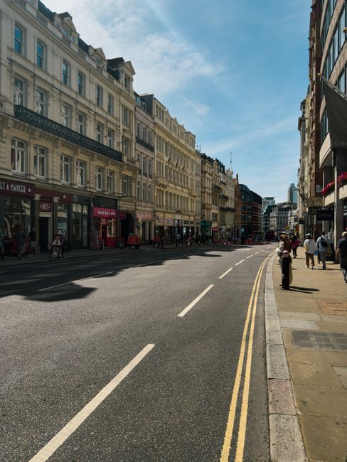 A wide view of Chiswick High Road in London, showing a row of elegant, multi-story Victorian and Edwardian style buildings with light-colored facades, large windows, and decorative architectural details. The street is lined with various retail shops and cafes on the ground floors, some with colorful signage and window displays. Pedestrians are walking along the sidewalk, and a few are waiting at a bus stop. The roadway has freshly painted white lines, double yellow lines along the curb, and appears clean and well-maintained with no visible litter or debris. Bright, natural daylight illuminates the scene under a partly cloudy blue sky, contributing to an overall atmosphere of vibrant urban activity. This image reflects a typical city street scene where regular surface cleaning and maintenance ensure hygiene and visual appeal, qualities that exemplify professional services like those offered by Chiswick Carpet Cleaning, as seen on the page titled 'Chiswick High Road carpet cleaning local service guide, CHISWICK' at chiswickcarpetcleaning.org.uk.