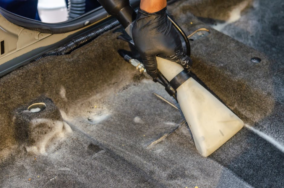 Close-up of a person wearing a black glove using a handheld cleaning device with a white spray head to perform surface cleaning on a dark, textured carpeted floor. The device appears to be emitting a spray or foam solution, indicating deep cleaning or sanitisation as part of professional carpet care. The surrounding area shows vacuum hoses and other equipment, suggesting a commercial or professional cleaning setting. The carpet has visible dust and dirt residue, and the cleaning process is highlighting the surface refresh and hygiene maintenance associated with residential or commercial carpet cleaning services offered by Chiswick Carpet Cleaning, tailored to the Gunnersbury Park area.