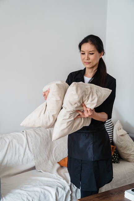 A woman is standing in a living room holding two beige cushions, positioned near a white sofa with a slightly wrinkled fabric cover. The room features a minimalist design with neutral-colored cushions, including one with black and white polka dots, and soft natural lighting. The woman has dark hair, wears a black apron over a white shirt, and appears engaged in domestic cleaning or cushion maintenance. The background shows a plain white wall, and the surface of the sofa and cushions is clean and well-maintained, reflecting hygiene and surface cleaning practices recommended for upholstery care. The setting suggests a focus on deep cleaning and maintaining pristine upholstery, aligning with services provided by Chiswick Carpet Cleaning for W4 homes, as highlighted in the Turnham Green upholstery cleaning tips page.