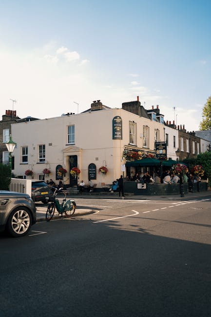 A street view of a charming, white-painted building housing a restaurant or pub, with large windows and flower baskets hanging outside, situated at a corner with outdoor seating on a paved patio. The surroundings include parked cars, a bicycle, and a lamppost, under a bright blue sky with a few clouds. The scene is well-lit and clean, highlighting the exterior surfaces and the sidewalk, indicative of regular maintenance and cleaning by Chiswick Carpet Cleaning to ensure hygiene and presenter appeal in commercial spaces. The area appears lively and inviting, with people visible near the entrance, and the overall setting emphasizes the importance of surface cleaning and upkeep in busy urban environments.