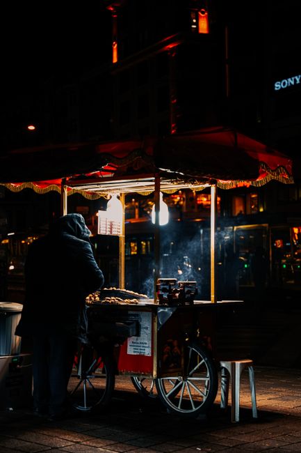 Nighttime street scene featuring a food cart under a canopy with red trim, illuminated by a bright overhead light, located in a commercial area with buildings and signage visible in the background. The vendor, dressed in dark clothing with a hood, is standing beside the cart, which is equipped with cooking appliances and utensils. The surrounding pavement appears clean and dry, with a stool positioned nearby. The scene reflects urban atmosphere and prepares for surface cleaning or sanitisation, which is a service offered by Chiswick Carpet Cleaning for maintaining hygiene in various environments, including commercial and residential spaces.