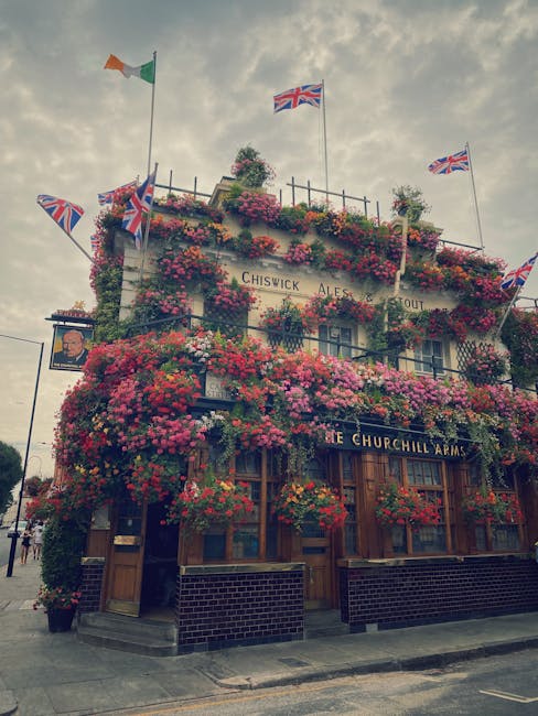 A street view of a charming, white-painted building housing a restaurant or pub, with large windows and flower baskets hanging outside, situated at a corner with outdoor seating on a paved patio. The surroundings include parked cars, a bicycle, and a lamppost, under a bright blue sky with a few clouds. The scene is well-lit and clean, highlighting the exterior surfaces and the sidewalk, indicative of regular maintenance and cleaning by Chiswick Carpet Cleaning to ensure hygiene and presenter appeal in commercial spaces. The area appears lively and inviting, with people visible near the entrance, and the overall setting emphasizes the importance of surface cleaning and upkeep in busy urban environments.